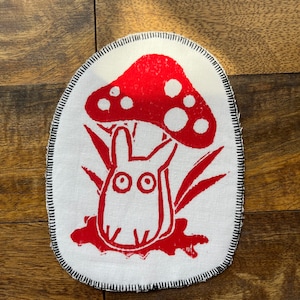 May include: A white fabric patch with a red mushroom and a white rabbit underneath. The mushroom has white spots and the rabbit has large eyes. The patch is surrounded by a black stitched border.