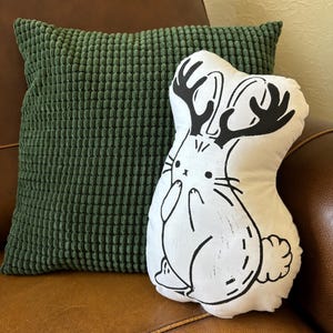 May include: A white pillow with a black and white illustration of a rabbit with antlers. The pillow is sitting on a brown leather couch next to a green pillow.