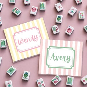 Puede incluir: Una vista en planta de fichas de Mahjong y dos tarjetas personalizadas. Las tarjetas tienen fondos a rayas, uno amarillo y blanco, el otro rosa y blanco. La tarjeta amarilla dice "Wendy" y la rosa dice "Avery". Las fichas son blancas con varios s&iacute;mbolos.