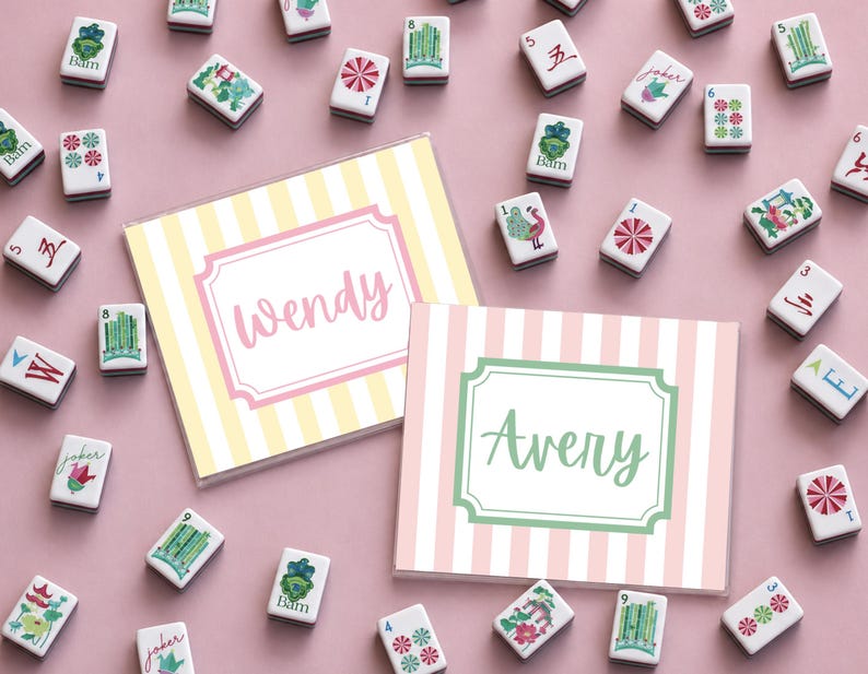 Puede incluir: Una vista en planta de fichas de Mahjong y dos tarjetas personalizadas. Las tarjetas tienen fondos a rayas, uno amarillo y blanco, el otro rosa y blanco. La tarjeta amarilla dice "Wendy" y la rosa dice "Avery". Las fichas son blancas con varios s&iacute;mbolos.