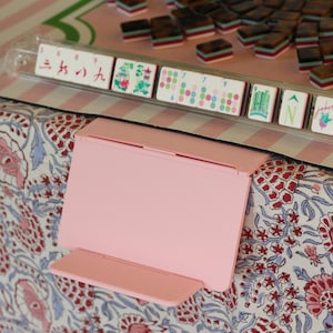 May include: A light pink plastic folding stand positioned in front of a Mahjong tile rack. The rack holds several white tiles with red, green, and blue symbols, numbers, and characters, including a tile marked with the word Joker in cursive script.