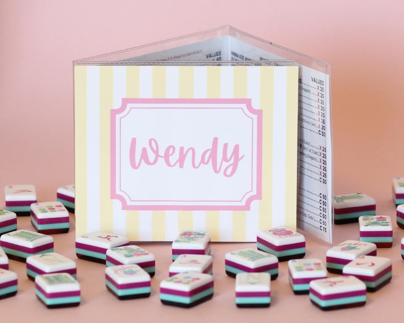 Puede incluir: Un letrero personalizado con el nombre "Wendy" en escritura rosa, sobre un fondo a rayas amarillas y blancas. Peque&ntilde;as piezas de juego coloridas con dise&ntilde;os florales est&aacute;n dispersas frente al letrero, creando una escena l&uacute;dica y decorativa.