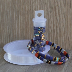 May include: A clear plastic container filled with small, colorful, rectangular beads. The container has a white lid with a handle. Next to the container is a spool of clear elastic thread and a bracelet made with the beads. The bracelet is multi-colored with blue, orange, white, black, and gold beads.