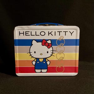 May include: A white metal Hello Kitty lunchbox with a blue handle. The front features Hello Kitty in blue overalls and a red bow, set against a rainbow striped background. Gold apple illustrations are on the right side.