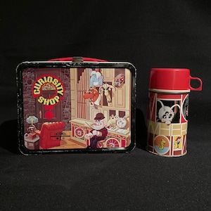 May include: Vintage metal lunchbox and thermos set. The lunchbox features a colorful illustration with the words "Curiosity Shop." The thermos has a red cup and a cartoon animal design. Both items are set against a black background.