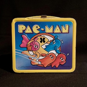 May include: Yellow metal Pac-Man lunchbox with a blue background and the Pac-Man logo. The lunchbox features a colorful graphic of Pac-Man and ghosts. The lunchbox has a metal clasp and a yellow handle.