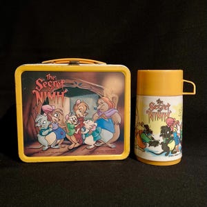 May include: Vintage yellow metal lunchbox and matching thermos featuring characters from "The Secret of NIMH." The lunchbox has a hinged lid and a handle. The thermos has a yellow lid and handle. Both items have a colorful illustration.