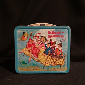 May include: Vintage blue metal lunchbox featuring a scene from Walt Disney's "Bedknobs and Broomsticks." The illustration shows characters on a bed, with a fish and bubbles. The lunchbox has a white handle and a metal clasp.