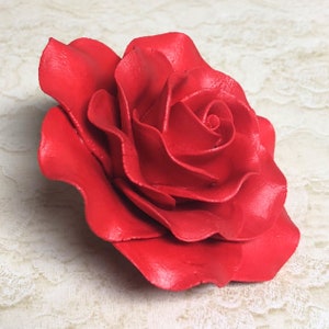 Pode incluir: Uma única rosa vermelha, grande e feita de tecido. A rosa tem um aspecto em camadas e com babados e é uma representação realista de uma rosa real.