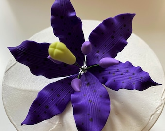 May include: A purple sugar flower with a yellow center and green stem. The flower has a textured surface and is made of fondant.