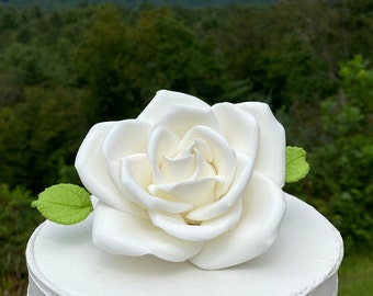 May include: A white fondant rose with green leaves sits atop a white cake. The cake is decorated with a rustic, textured finish.
