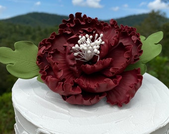 May include: A burgundy flower cake topper with ruffled petals and white accents, on a white frosted cake. Green leaves are on either side of the flower. The cake is set against a backdrop of mountains and a blue sky.