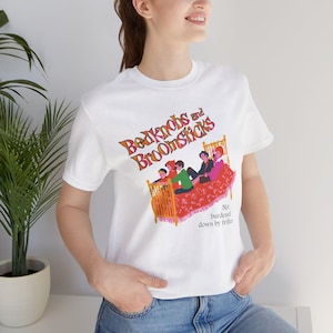 May include: A white short-sleeve t-shirt featuring a graphic of a bed with four figures and the text Bedknobs and Broomsticks and Not burdened down by frillies. The design uses orange, red, green, and pink colors to depict the scene.