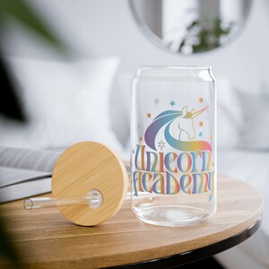 Puede incluir: Un vaso de vidrio transparente en forma de lata con tapa y pajita de madera. El vaso presenta un diseño de unicornio colorido con las palabras "Unicorn Academy". El diseño incluye estrellas y una melena arcoíris.