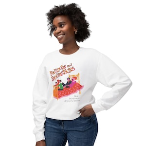 May include: A white long-sleeve sweatshirt featuring a graphic print of the text Bedknobs and Broomsticks above an illustration of four people sitting on a bed with a red patterned quilt and the text Not burdened down by frillies.