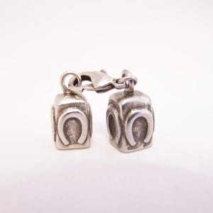 May include: Two silver-toned metal charms, each featuring a raised horseshoe emblem on a square-shaped base. The charms are connected by a small silver-toned lobster claw clasp, creating a linked pair of decorative jewelry components.
