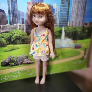 May include: A doll with long auburn hair wearing a yellow top with colorful designs and white shorts. The doll is standing in front of a backdrop of a city skyline and a park.