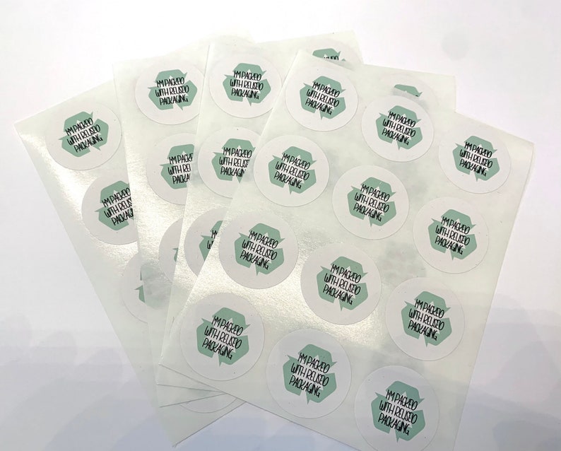 May include: A stack of white sheets with multiple round stickers that say "I'm packed with reused packaging". The stickers have a green recycling symbol with a green background.
