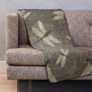 May include: A throw blanket draped over a sofa, featuring a repeating pattern of stylized dragonflies. The dragonflies have cream-colored wings and blue accents, set against a textured, gray-brown background. The sofa is a neutral gray color.