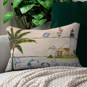 May include: Rectangular decorative pillow with a coastal theme. The pillow features a palm tree, lighthouse, seahorse, and compass rose design in shades of green, blue, and beige. The pillow is placed on a light gray sofa with a cream-colored blanket.