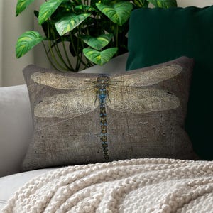 May include: Rectangular decorative pillow featuring a detailed dragonfly illustration. The pillow has a textured, neutral-toned background with a large dragonfly in shades of beige, blue, and gold. The pillow is placed on a gray sofa with a white blanket.
