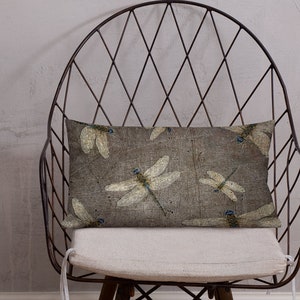 May include: A decorative throw pillow with a beige background and a pattern of delicate dragonfly illustrations in shades of brown, white, and blue. The pillow is sitting on a black metal chair with a white cushion.