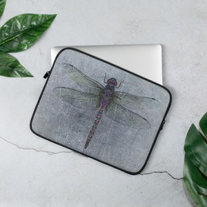 May include: A laptop sleeve with a dragonfly design. The dragonfly is a vibrant purple, blue, and green on a gray background with a distressed texture.