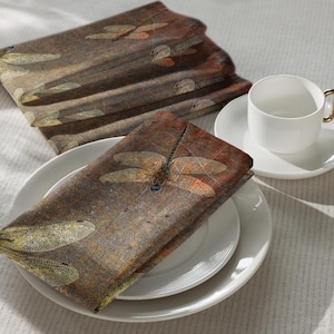 May include: Set of six brown and beige fabric napkins with a dragonfly design. The napkins feature a distressed, vintage look with a subtle metallic sheen.