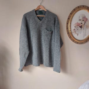 May include: A gray v-neck sweater with a patch pocket on the left side. The sweater is hanging on a wooden hanger against a light beige wall.