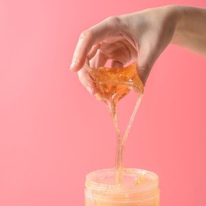 May include: A hand holding a golden brown sugar wax, dripping into a clear jar. The wax is thick and gooey, and the jar is filled with more of the same wax.