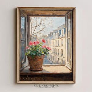 Op de afbeelding: Een olieverfschilderij toont een venster met een terracotta pot met roze rozen op een houten vensterbank. Het open raam onthult een Europese straatscène met gebouwen en een blauwe lucht. Het schilderij is ingelijst in een goudkleurige houten lijst.