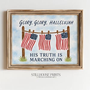 May include: A framed wall art print featuring a watercolour illustration of three American flags hanging on a clothesline between two wooden posts. The text GLORY, GLORY, HALLELUJAH and HIS TRUTH IS MARCHING ON is printed in dark blue against a light blue sky.