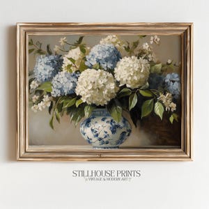 Op de afbeelding: Een ingelijste bloemenprint met een vaas vol witte en blauwe hortensia's. De vaas heeft een blauw en wit bloemmotief. Het kunstwerk is gelabeld "STILLHOUSE PRINTS VINTAGE & MODERN ART". De lijst is lichtbruin.