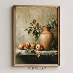Könnte beinhalten: Ein gerahmtes Stillleben mit einem terrakottafarbenen Keramikkrug neben drei roten und grünen Äpfeln auf einem Steinsims. Ein belaubter Zweig ragt aus dem Krug hervor. Der Text Stillhouse Prints Vintage & Modern Art ist zu sehen.