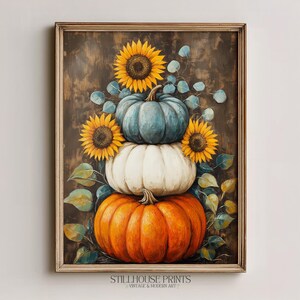 Könnte beinhalten: Ein gerahmter Kunstdruck mit einem vertikalen Stapel aus drei Kürbissen in Blau, Weiß und Orange. Das Arrangement ist mit leuchtend gelben Sonnenblumen und grünen Eukalyptusblättern vor einem dunklen, strukturierten, braunen Hintergrund akzentuiert.