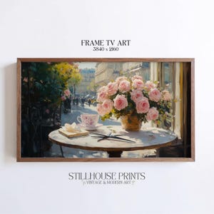 May include: A framed art print depicts a Parisian cafe scene. A round table holds a teacup, a book, and a vase of pink roses. The artwork is labeled "FRAME TV ART" and "STILLHOUSE PRINTS VINTAGE & MODERN ART."