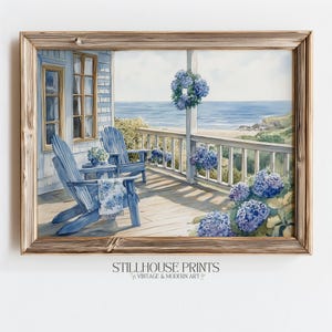 Könnte beinhalten: Aquarellmalerei einer Küstenveranda-Szene mit zwei blauen Adirondack-Stühlen, einem Tisch und einer Decke. Die Veranda blickt auf das Meer, mit blauen Hortensien und einem Kranz. Das Kunstwerk ist in einem Holzrahmen gerahmt.
