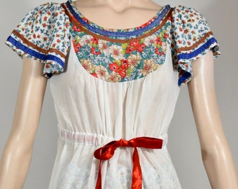 Puede incluir: Una blusa blanca de campesina con un cuello de encaje y un lazo de cinta roja. La blusa se combina con una falda blanca con estampado floral y ribete rojo.