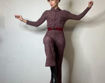 May include: A long-sleeved, high-neck jumpsuit in a textured, heathered maroon knit fabric. The garment features a front zipper closure, a small chest pocket, and a wide, solid dark red belt with a large, circular, decorative buckle.