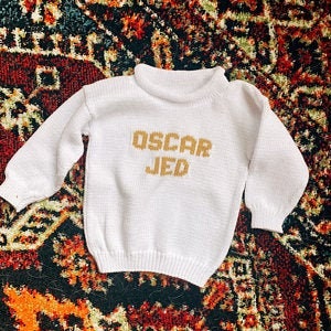 May include: White knitted sweater with a gold knitted inscription that reads "OSCAR JED".