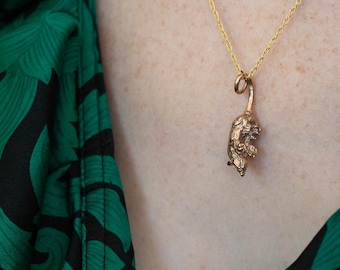 May include: A gold chain necklace with a small, detailed, gold animal pendant. The necklace is worn by a person wearing a green and black patterned blouse.