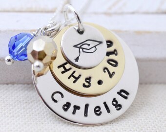 May include: A silver necklace with a layered charm featuring a graduation cap, the text "HHS 2019", and the name "Carleigh". A blue crystal bead is attached to the chain.