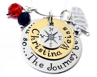 May include: A silver necklace with a compass charm, two layered discs with the text "The Journey Begins... Christina Weiss 2018" and two red and blue crystal beads.