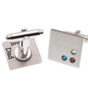 May include: Silver cufflinks with a square design. One cufflink has the inscription "Dad" and the other has three small gemstones.