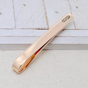 May include: A rose gold tie bar with a brushed finish. The tie clip features a small, black infinity symbol design on one end. The tie bar is shown on a white wooden surface.