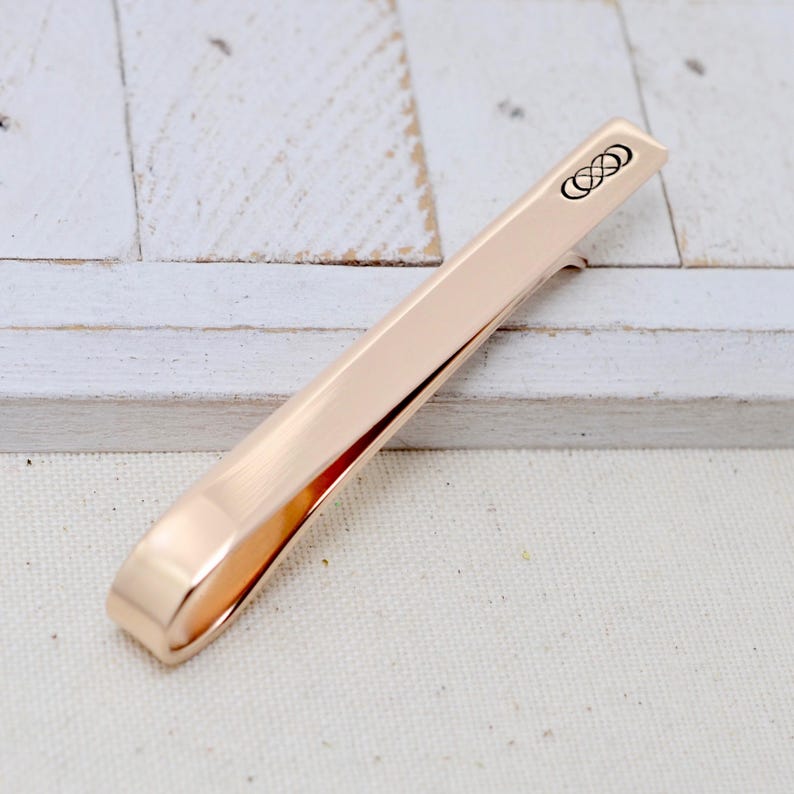 May include: A rose gold tie bar with a brushed finish. The tie clip features a small, black infinity symbol design on one end. The tie bar is shown on a white wooden surface.