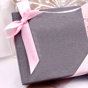 May include: A gray gift box with a pink satin ribbon tied in a bow. The box is wrapped in a pink ribbon.