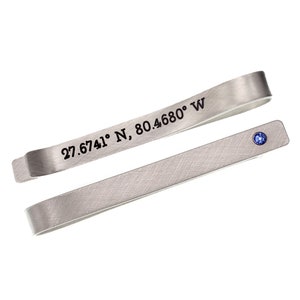 May include: Silver tie bar with a blue gemstone and engraved coordinates: 27.6741° N, 80.4680° W.