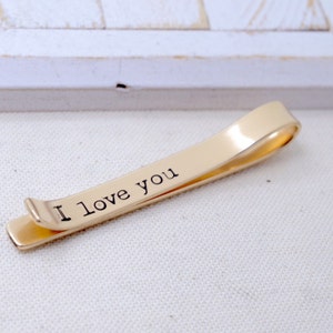 May include: A gold-colored tie clip with the words "I love you" printed in black. The clip is made of a shiny metal and has a simple, elegant design. The tie clip is a classic accessory.