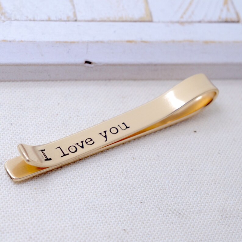 May include: A gold-colored tie clip with the words "I love you" printed in black. The clip is made of a shiny metal and has a simple, elegant design. The tie clip is a classic accessory.